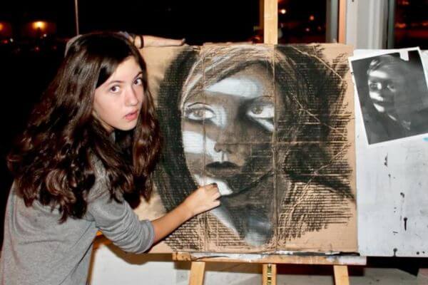 A student with dark hair holding charcoal, creating artwork on cardboard