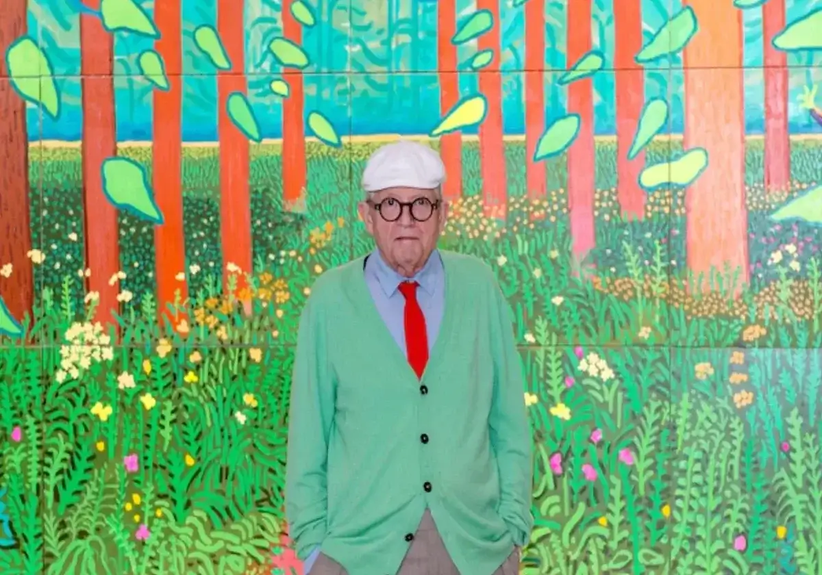 Man in Green sweater and red tie with white hat stands in front of brightly colored artwork